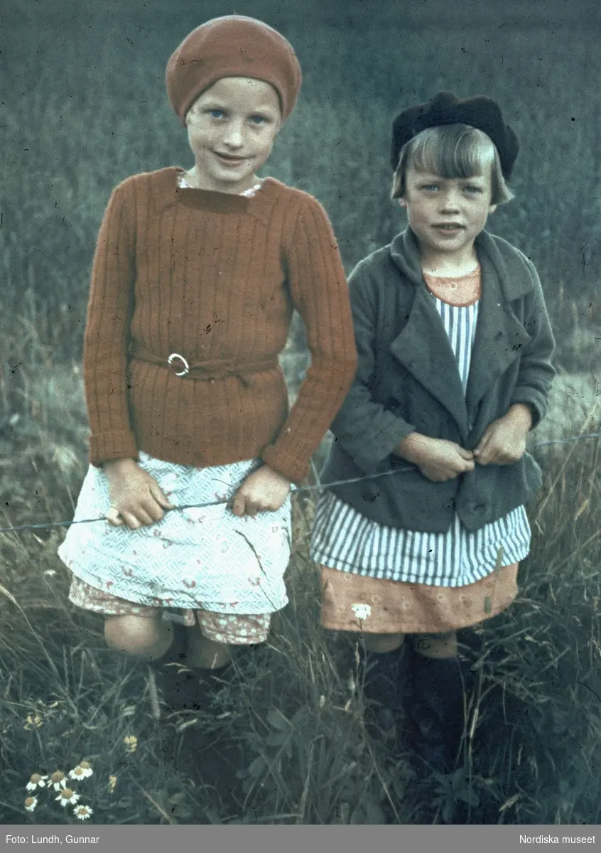 Statarbarn på trettiotalet. Två flickor med varsin basker och stövlar står i högt gräs.