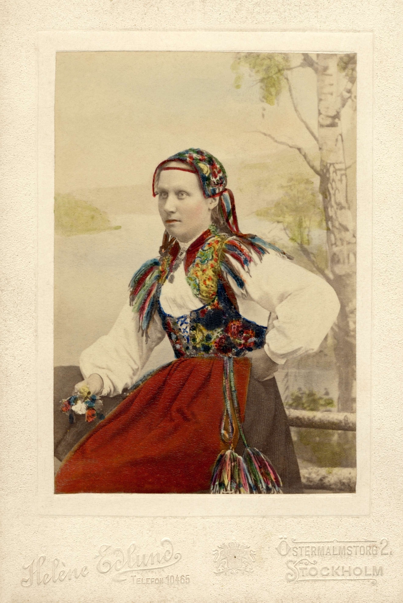 En ung kvinna poserar i en folkdräkt med schalen instoppad i västen. Handkolorerat fotografi.