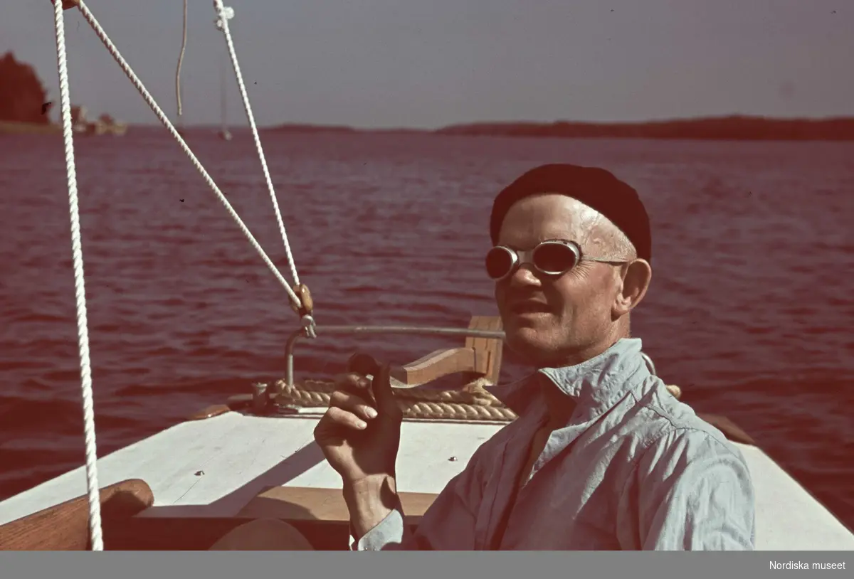 En man i svart basker och solglasögon sitter till rors i en segelbåt.