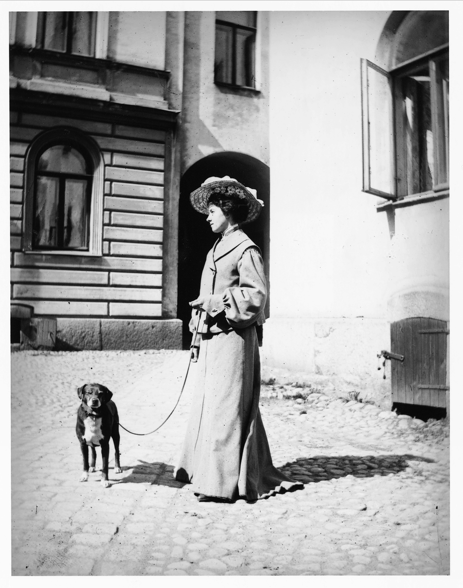 Kvinna på promenad med hund i Stockholm, 1890-1910. De har stannat framför några byggnader. Kvinnan är klädd i dräkt med vida ärmar och lång kjol, samt vidbrättad stråhatt. Hon håller hunden i koppel. 