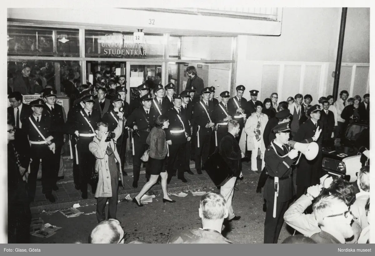 Kårhusockupationen vid Stockholms universitet i slutet av maj 1968. Kvällsbild från Studentkårens entré. Utanför har poliser barrikaderat dörrarna. En polisman talar till folksamlingen i megafon. En pressfotograf tar en bild.