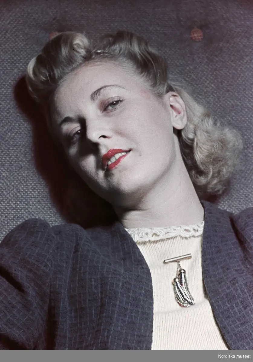 Porträtt av blond kvinna med rött läppstift, stickad blus med en brosch.