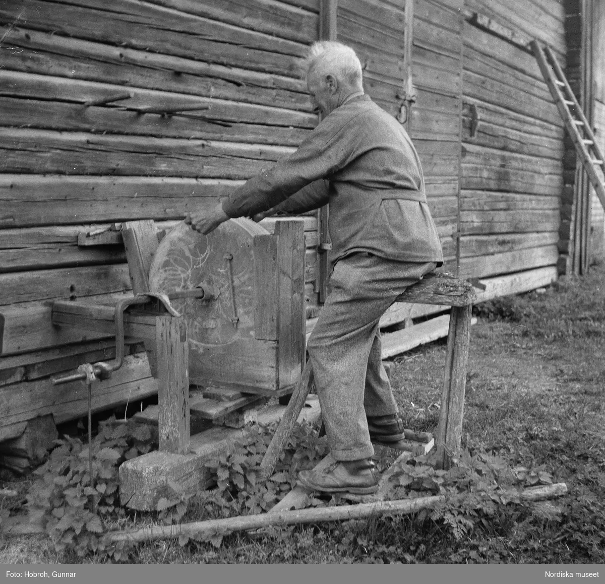 Osmundsbergs by i Boda socken, Dalarna. Slipsten, konstruerad av ägaren, Röjås Erik Andersson, vilken själv sitter på sätet. Den drivs med båda fötterna genom trampning vid ändarna av en bräda. Stenen är från Orsa.
