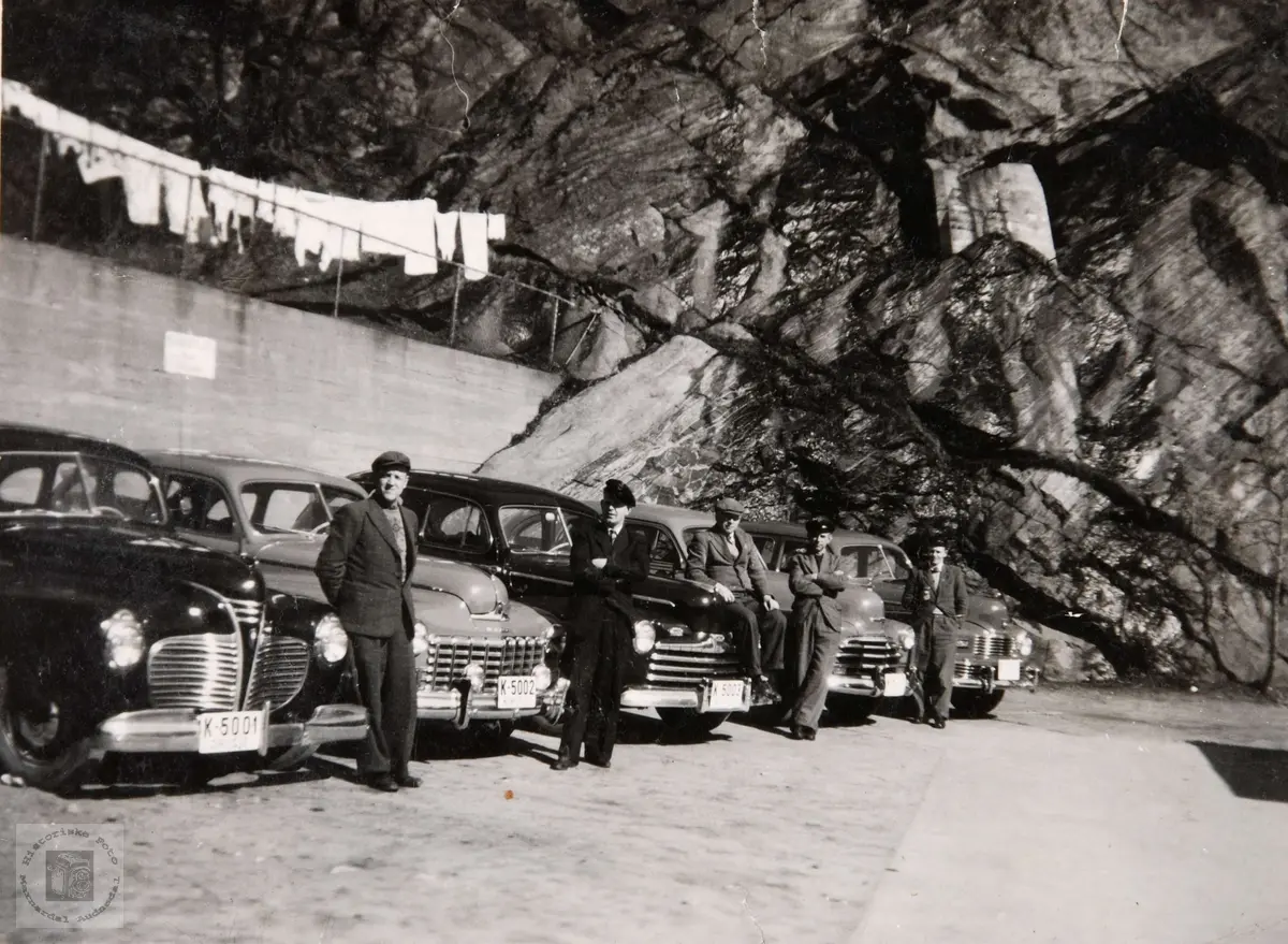 Drosjeparken i Mandal på 1950 tallet.
Drosjene fra venstre: K-5001 Plymouth 1940, K-5002 Dodge 1946-48, K-5003 Ford 1946, Chevrolet 1947, Nash 1946-48. 
Brukerkommentar september 2020: Til venstre: Rolf Morris Jahnsen.