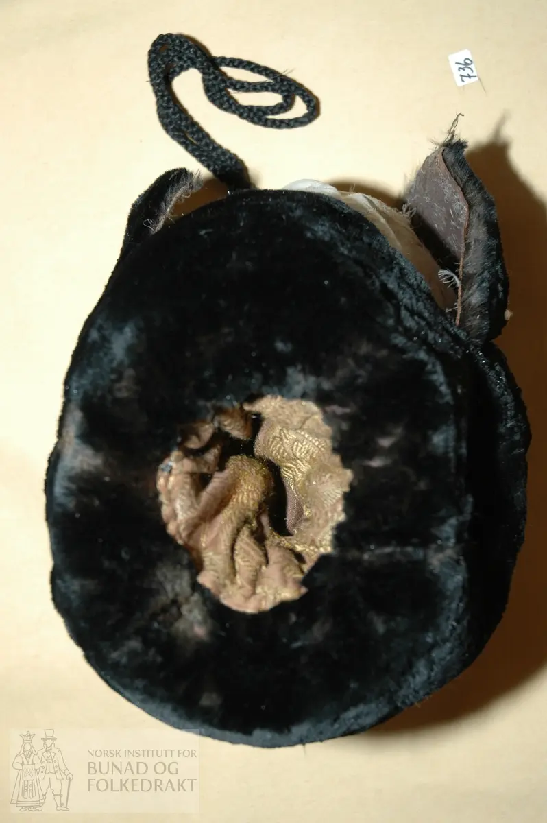 Muffe sydd av svart kaninskinn fora med mønstra damask i brunt og gyllent. Fyll av dun i ubleika bomullslerret.