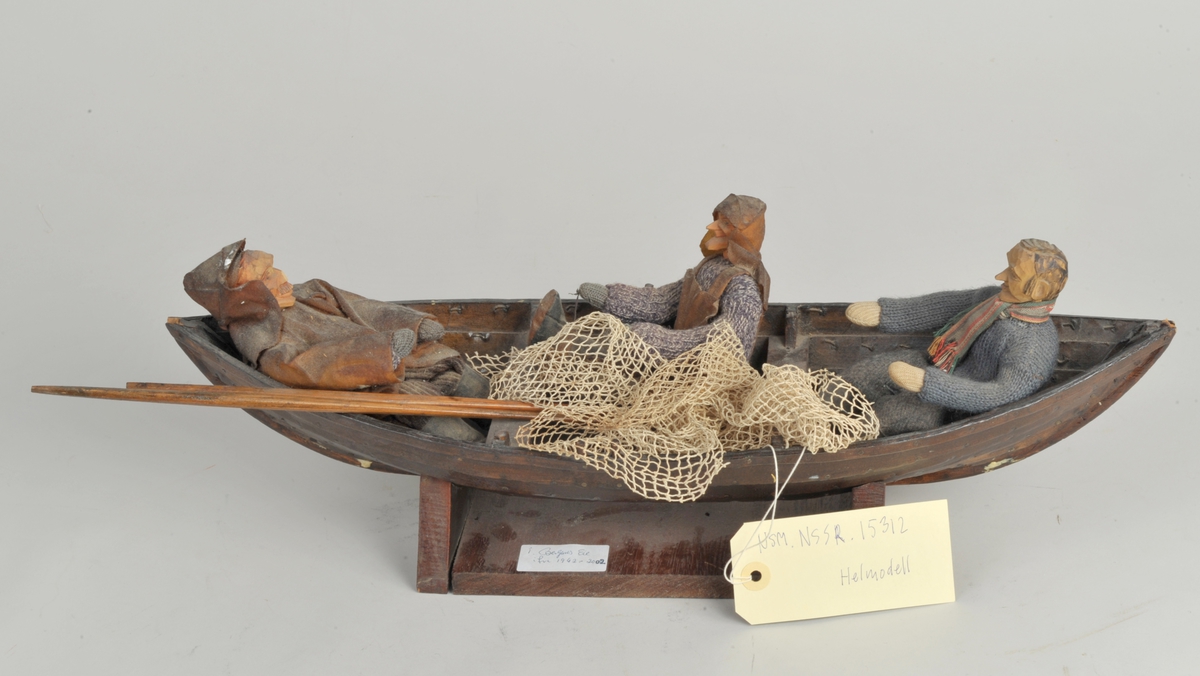 Modell av robåt, grovt tillaget. Tre figurer sitter oppi båten. Båten står på en løs sokkel.
