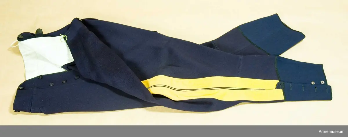 Mörkblå klädesdiagonal med dubbla klädeslister och emellan dem en gul passpoal. 6 knappar av svart bakelit i gylfen.  Byxbenen slutar strax under vaden där de knäpps med resp. 5 knappar av pärlemor. 
Har för ridbyxor s.k. vid tillskärning.
Har tillhört Gustav VI Adolf (1882-1973).