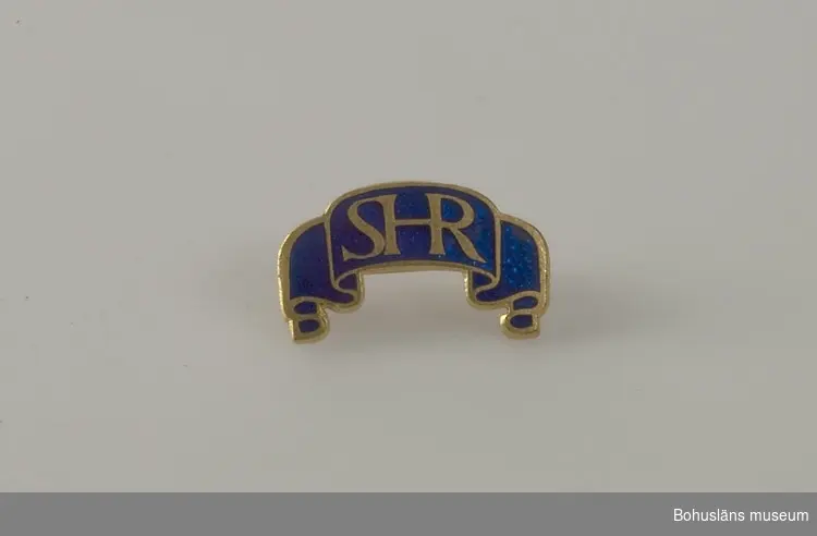 Brosch i form av ett veckat band med bokstäverna "SHR" i mitten.
Guldfärgat mot blå emalj. 
Nål på baksidan för att fästa broschen.