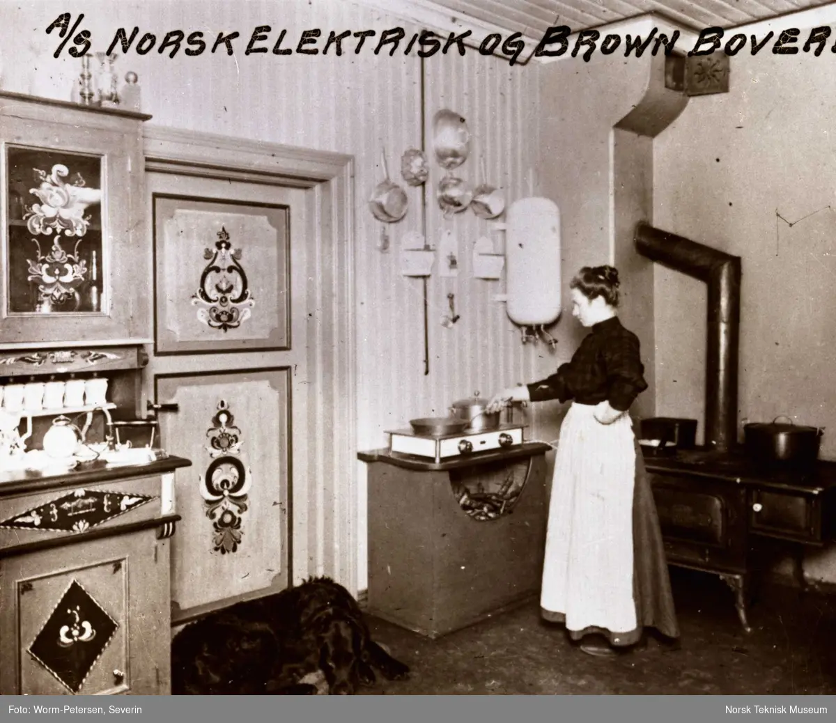 Reklameserie fra A/S Norsk Elektrisk Brown Boveri (NEBB): Kjøkkeninteriør fra Heje i Hallingdal med elektrisk kokeplate, varmtvannsbeholder og vedkomfyr i bakgrunnen. Publisert i Landbruk og elektrisitet 1914, s. 64.