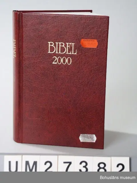 Bibel inbuden i rött konstläderband med texten "BIBEL 2000" i nedsänkt guldrelief på framsida och rygg. 
Bibelns text är  i nyöversättning, den tredje med officiell karaktär. Huvudsyftet är att vara huvudtext för bibeln inom det svenska språkområdet. De båda tidigare översättningarna utgavs 1541 och 1917. Bibelns grundtexter skall här översättas till nutida svenskt språk.
Bibekommissionen tillsattes 1972 och avslutar sitt arbete i och med utgången av år 2000. Texten i denna bibel godkändes av Bibelkommissionens styrelse 1999.
Avslutande historisk presentation i fyrfärg av bibelns originalspråk, skriftspråk,  tidsaxlar,  karta, arkitektur, högtider, betydelsefulla personer i Gamla och Nya testamentet, vardagsliv i bibels länder, innehållsöversikt m.m.

Text på försättsbladet:
"BIBEL 2000 
GAMLA TESTAMENTET
APOKRYFERNA
NYA TESTAMENTET
Bibelkommissionens översättning 
Marcus Förlag"

Inköpt  för 149 kronor, extrapris.

För information om Millennieinsamlingen, se UM27360.
