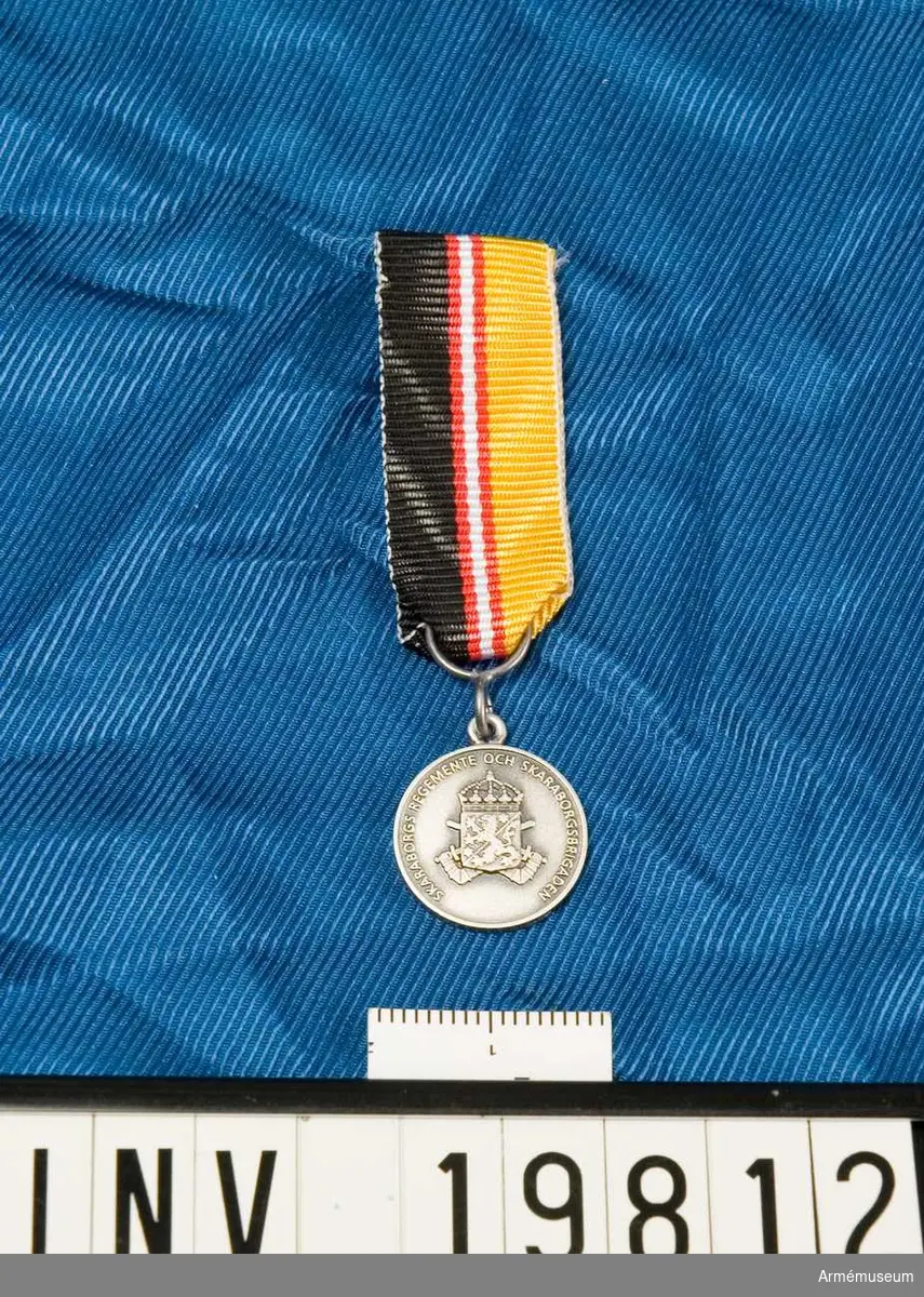 Medaljen är rund. Vapenskölden lagd över två korslagda bepansrade armar hållande varsitt svärd och krönt med en kunglig krona. I skölden ett lejon åtföljt av två stjärnor. På frånsidan en lagerkrans. Inskription: Skaraborgs regemente och Skaraborgsbrigaden. Band kluvet i svart och gult med en vit rand i mitten åtföljd av en röd rand på vardera sidan. Miniatyrmedaljen förvaras i ask tillsammans med en medalj.