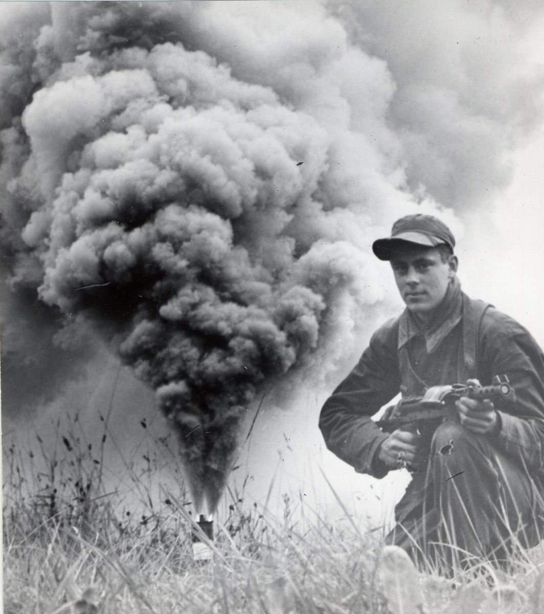 Soldat står på knä med eldberedd kulsprutepistol m/1945 i händerna. I bakgrungen brinner en rökfackla.