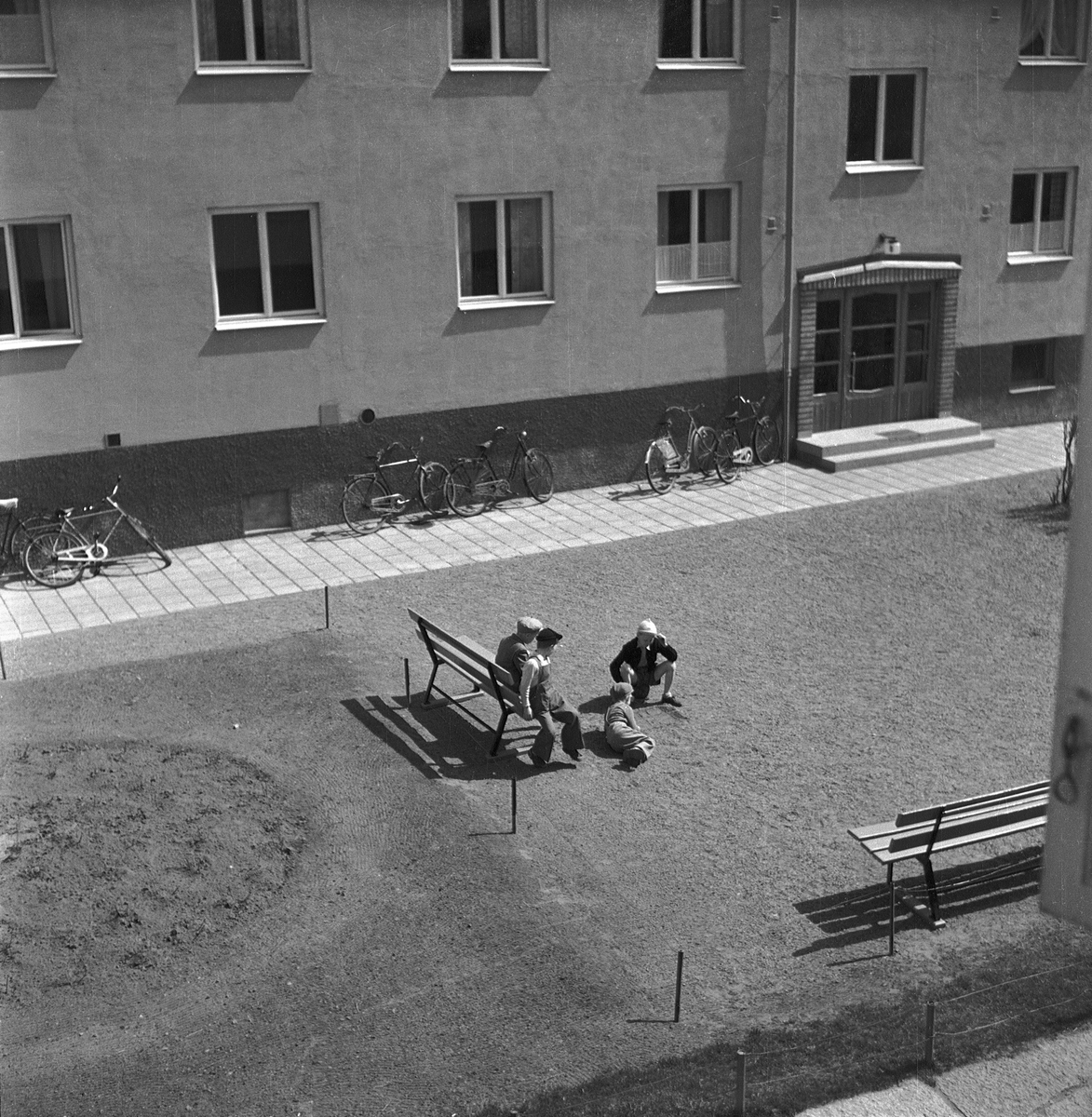 Fyra barn på en gård vid ett hyreshus i Jönköping. Två av dem spelar luffarschack på grusplanen och två sitter på soffan intill och tittar på. Några cyklar står lutade mot husväggen.