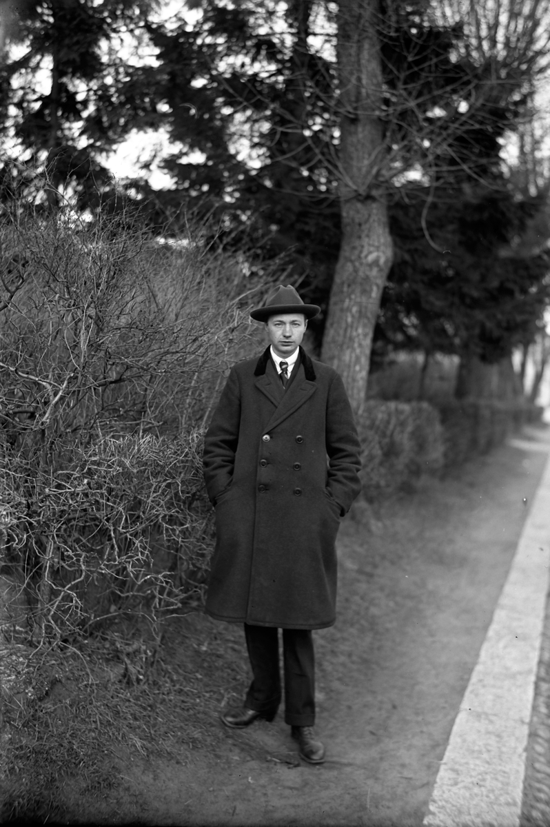 Gustav Andersson klädd i rock och hatt, står vid en vägkant. Bredvid honom växer en avlövad buske och i bakgrunden syns träd och en häck.
