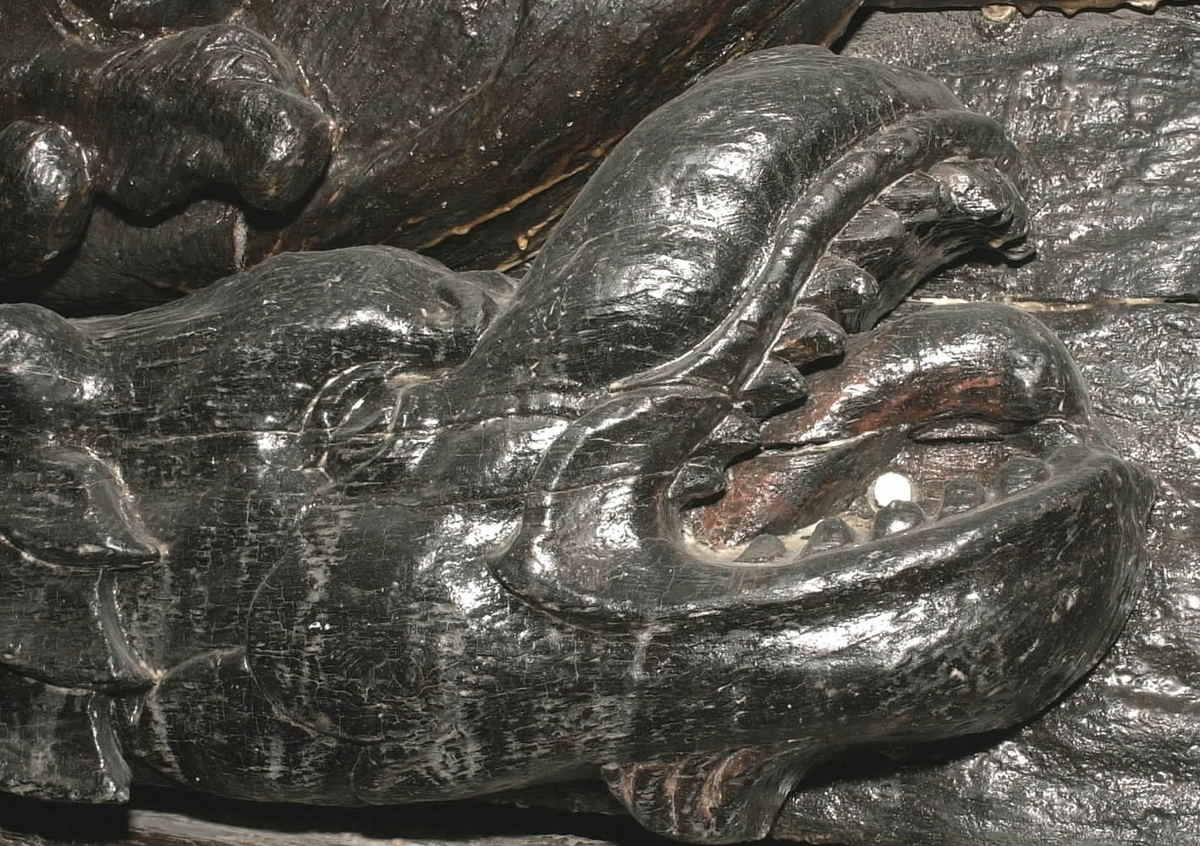 Skulptur i form av en drake (delfin?), återgiven i höger profil.
Draken har ett stort huvud med uppåtvriden nos och vidöppen käft, blottande en tjock tunga och spetsiga tänder. Halvrundade vingar. Stjärten fortsätter uppåt utmed stjärten till en triton, se fyndnummer 00020. Baksidan är slät.
Skulpturen är mycket välbevarad.

Text in English: A sculpture of a dragon or dolphin in right profile.
The dragon has a large head with upturned nose and wide-open mouth, exposing a thick tongue and pointed teeth. Semi-round wings. The tail extends upwards with the tail to a Triton, see No. 00020. The back is smooth.
The sculpture is well preserved.
