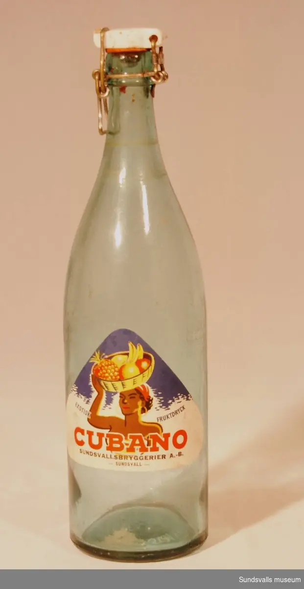 En läskedrycksflaska med patentkork från Sundsvallsbryggerier AB. Flaskan är tillverkad i blåtonat glas och är försedd med en trekantig etikett med texten 'EXOTISK FRUKTDRYCK CUBANO, SUNDSVALLSBRYGGERIER A.-B. - SUNDSVALL -'.