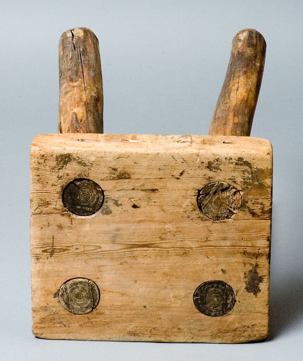 Pall, handgjord av trä, furu. Fyrkantig sits med fyra genomgående itappade ben. 
Skrivet under sitsen: Skansens rekvisita (Delsbogården) BOLLNÄS.
