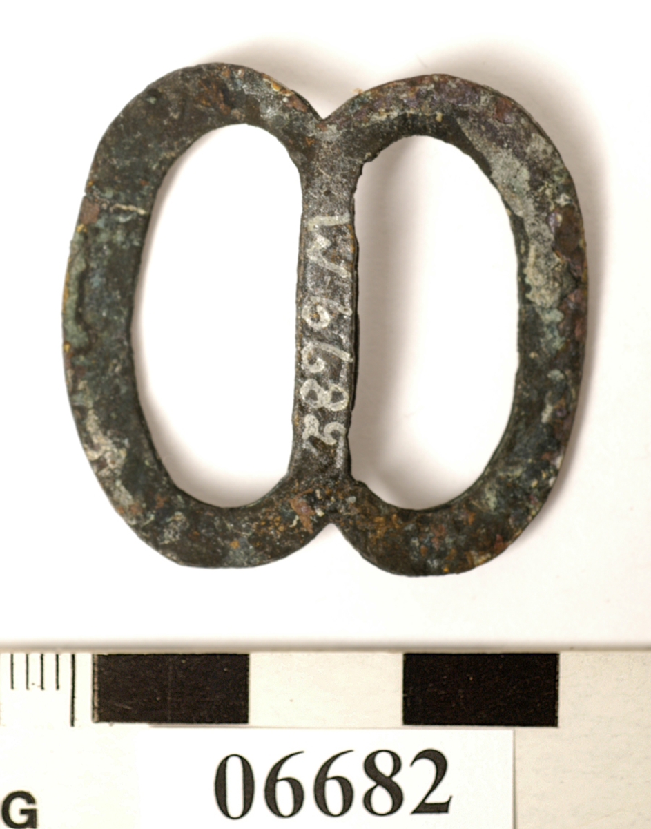 Sölja. Spänne. Formad som en åtta. Lätt böjd på mitten.

Text in English: One metal buckle.
One side of metal (probably front) is convex, the other side (probably back) is flat, with defined edges.
The whole piece is concave, the center bar sits lower than the sides.  When viewed from the side, it resembles a wide V.  When viewed from the front or the back, it resembles a flattened figure 8.
Handmade out of what appears to be one piece of metal, two solder points visible.