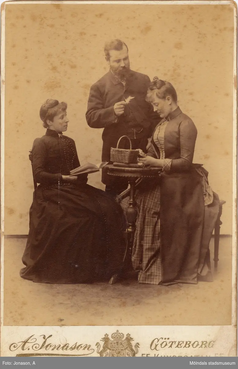 Ateljébild med två damer sittandes vid bord och en herre som står bakom hållandes en fjäder i handen. Mannen är Martin Stenström, som ägde godset Kristinedal och firman Buhrmans änka, damkollektion. Kvinnan till höger är Kerstin Stenström (född Svanfeldt), kvinnan till vänster är okänd.