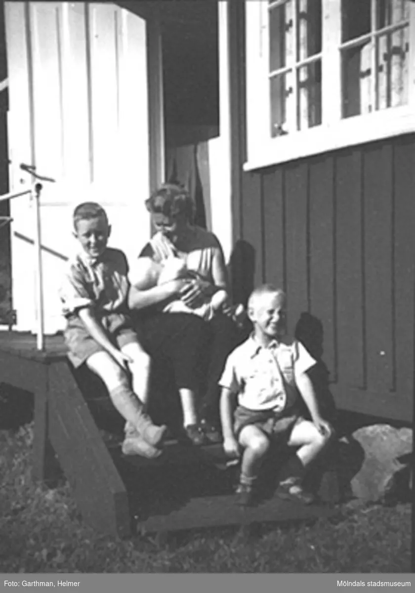 Astrid Garthman sitter på trappan med yngste sonen Jerry i famnen. På hennes högra sida sitter Leif och till vänster sitter Alf. Familjen Garthman lånade 1955 en sommarstuga av Artur Rolén som var målare på API. Stugan låg i Stannum (Lerum) nedanför sjön Mjörn.