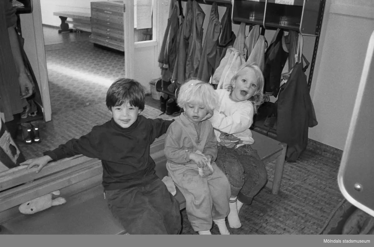 Dagisbarnen Daniel, Rebecka och Malin sitter på en träbänk inomhus, i entrén till Hoppetossan på Katrinebergs daghem. I bakgrunden ser man barnens bås med deras ombytes- och regnkläder.
