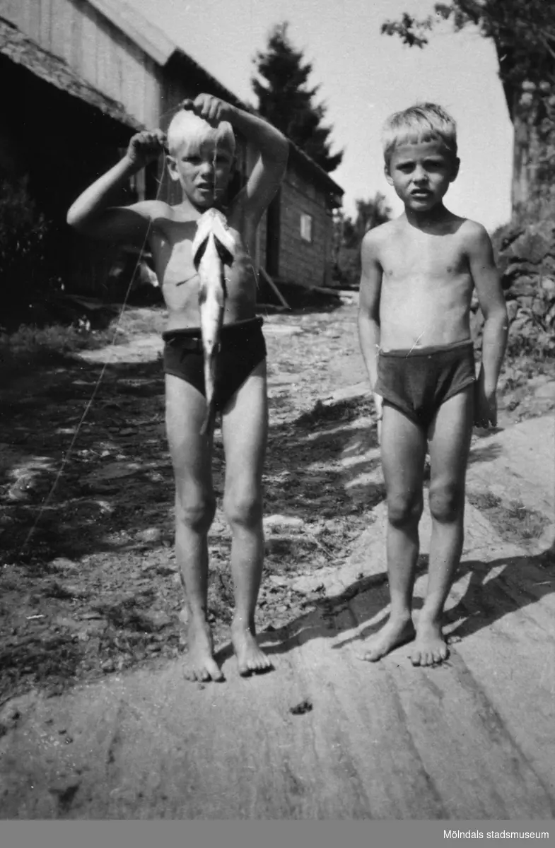 Två yngre pojkar som har fiskat, troligtvis i Tulebosjön cirka 1930. Pojken till vänster håller ett spö med en fångad fisk på kroken. I bakgrunden ses en väg, byggnader och träd.