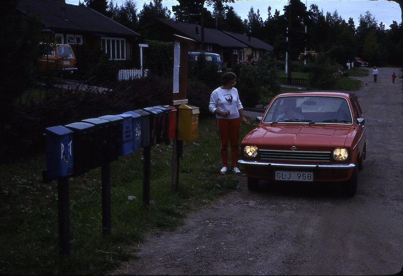 Lantbrevbärare Mikael Mattsson har kommit till Rocksta. Gunvor Wessberg står vid bilen och samtalar med honom. I förgrunden syns en samling postlådor och en gul brevlåda.