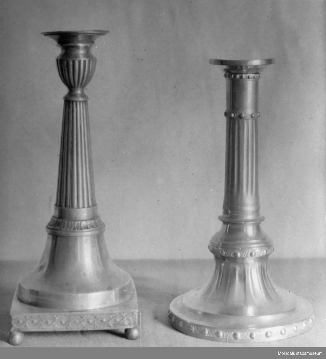 Två ljusstakar av metall med varierande höjd och design, ståendes på var sin sockel. Gunnebo slott 1930-tal.