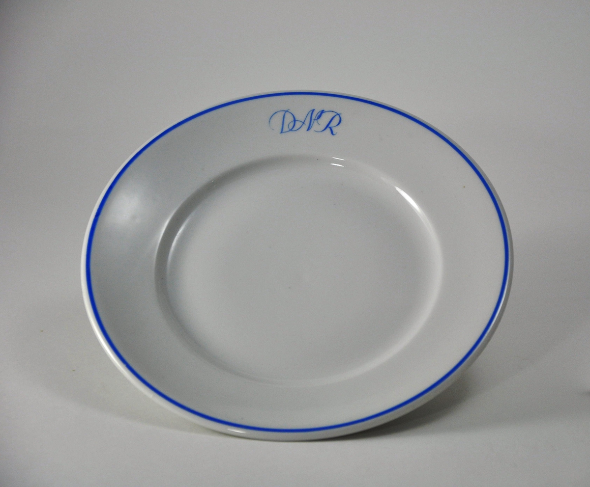 Flat tallerken i hvit porselen. A/S Den Norske Reiseeffektfabriks initialer "DNR" og munningsrand er påmalt i blått.