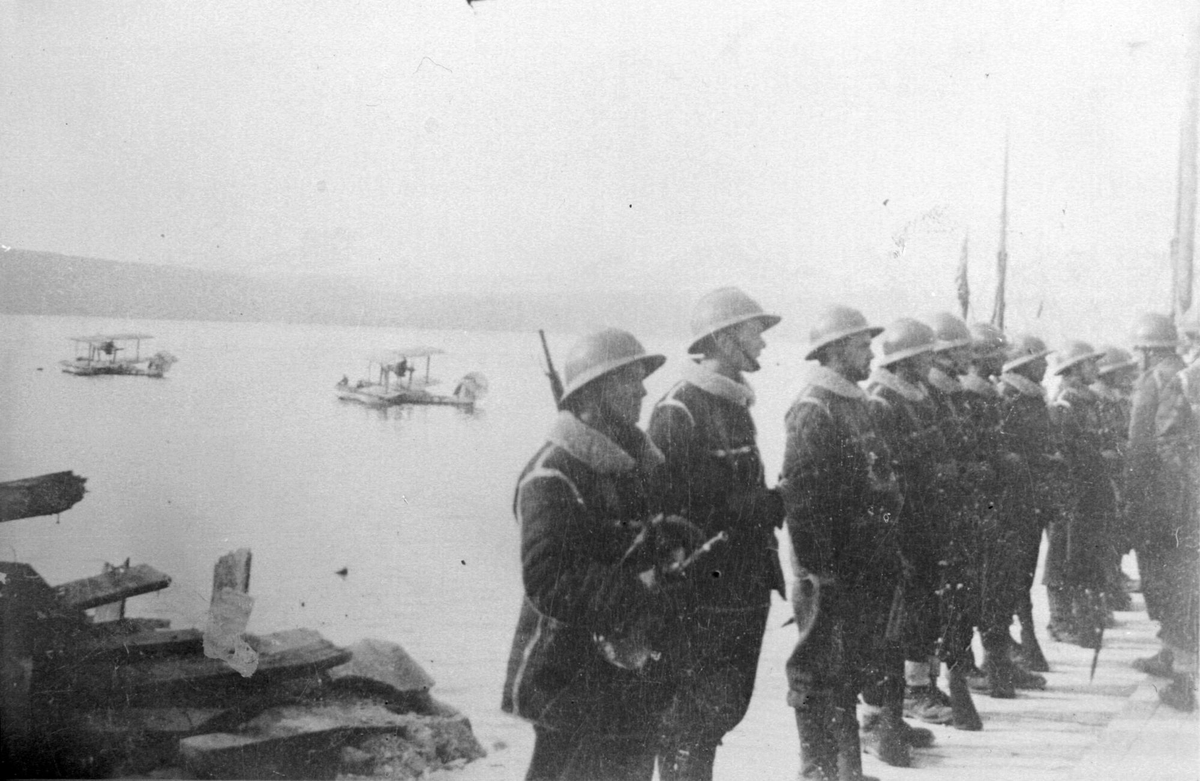 Franske soldater oppstilt på dampskipskaia. To britiske sjøfly ligger på sjøen i bakgrunnen.
