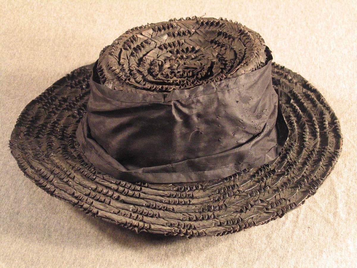 Breibremma hatt med pull med søkk. Rundt pullen er eit breitt silkestykke med mange merke etter hattenål på eine sida. Fôr i pullen.