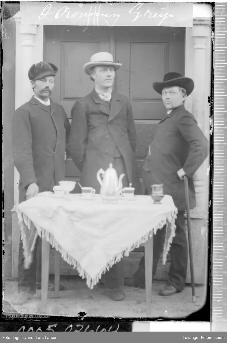 Gruppebilde av tre menn ved et bord foran et inngangsparti.