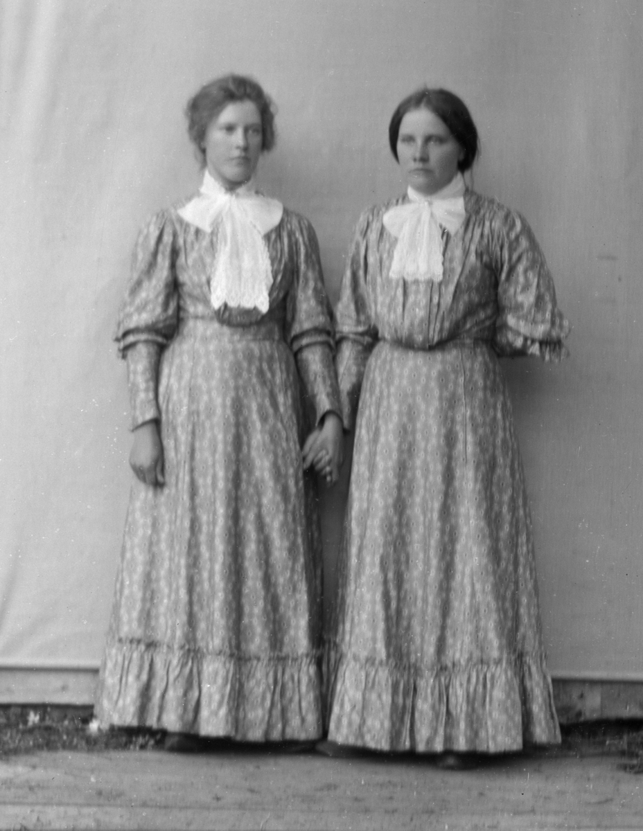 To kvinner i helfigur kledd i mønstrete kjoler, stående foran hvitt lerret på husvegg