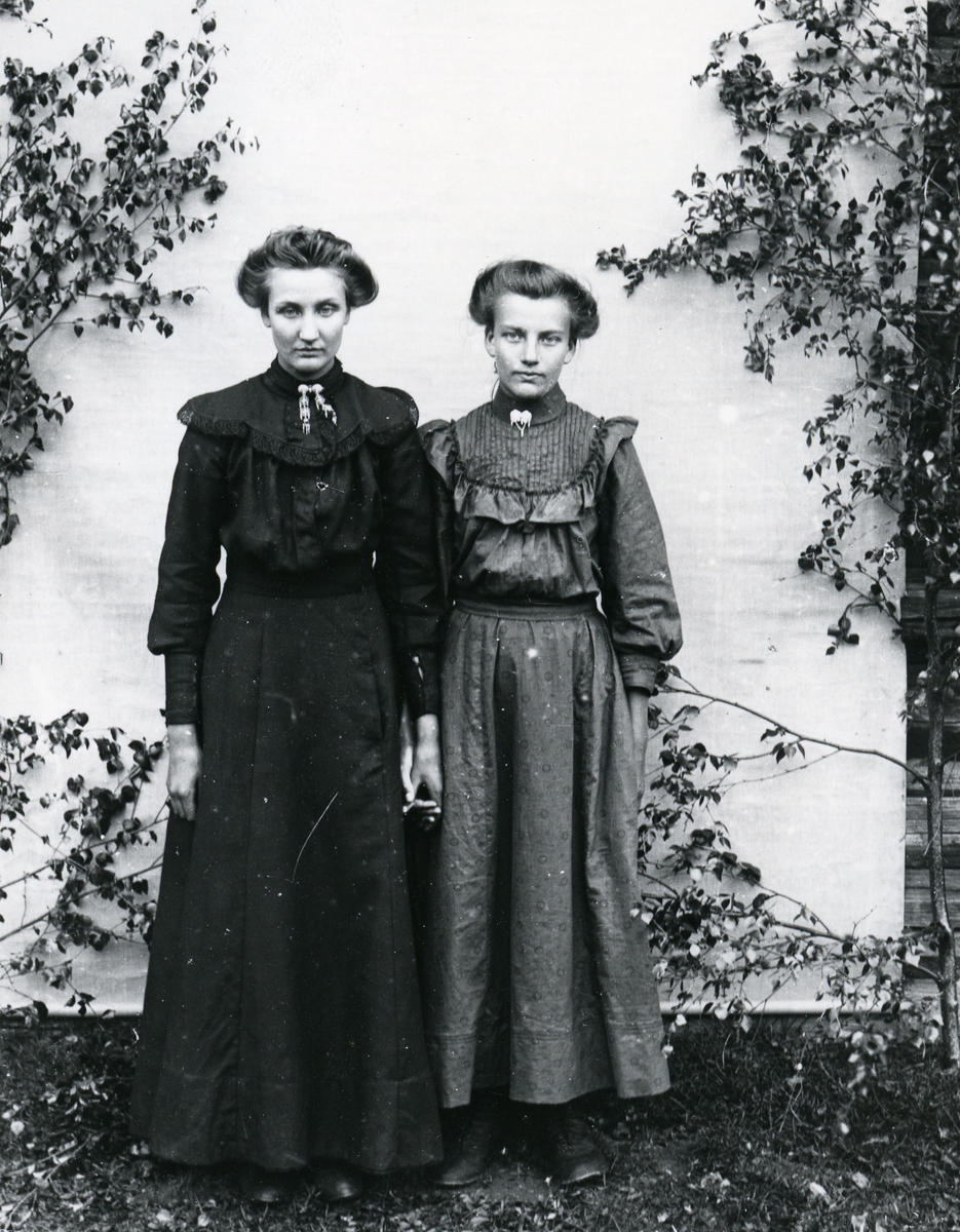 To kvinner i helfigur, hånd i hånd, stående foran lerret, med bjørkekvister