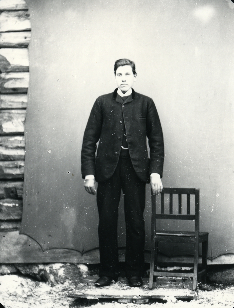 Mann i helfigur, stående ved stol, foran lerret på tømmervegg