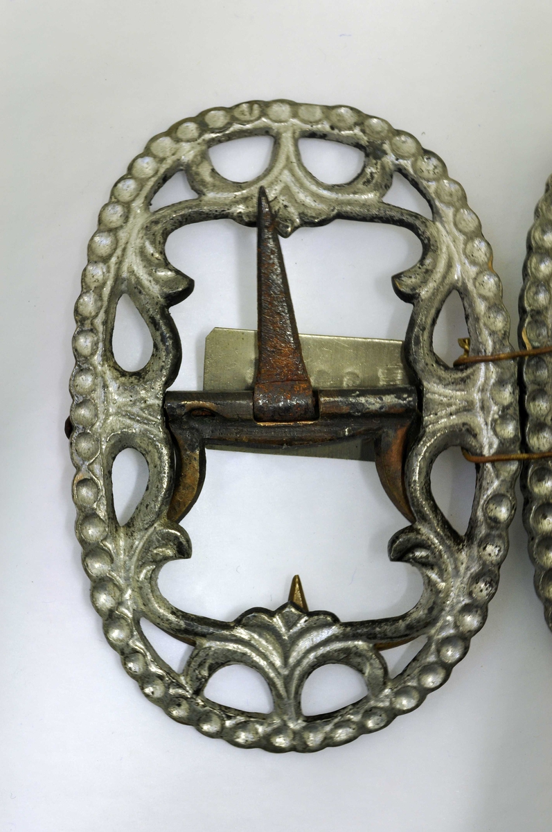 Fra protokoll: 1 par skospænder av fortinnet bronse med gjennembrutt bladverk og rand av hul perlesnor.