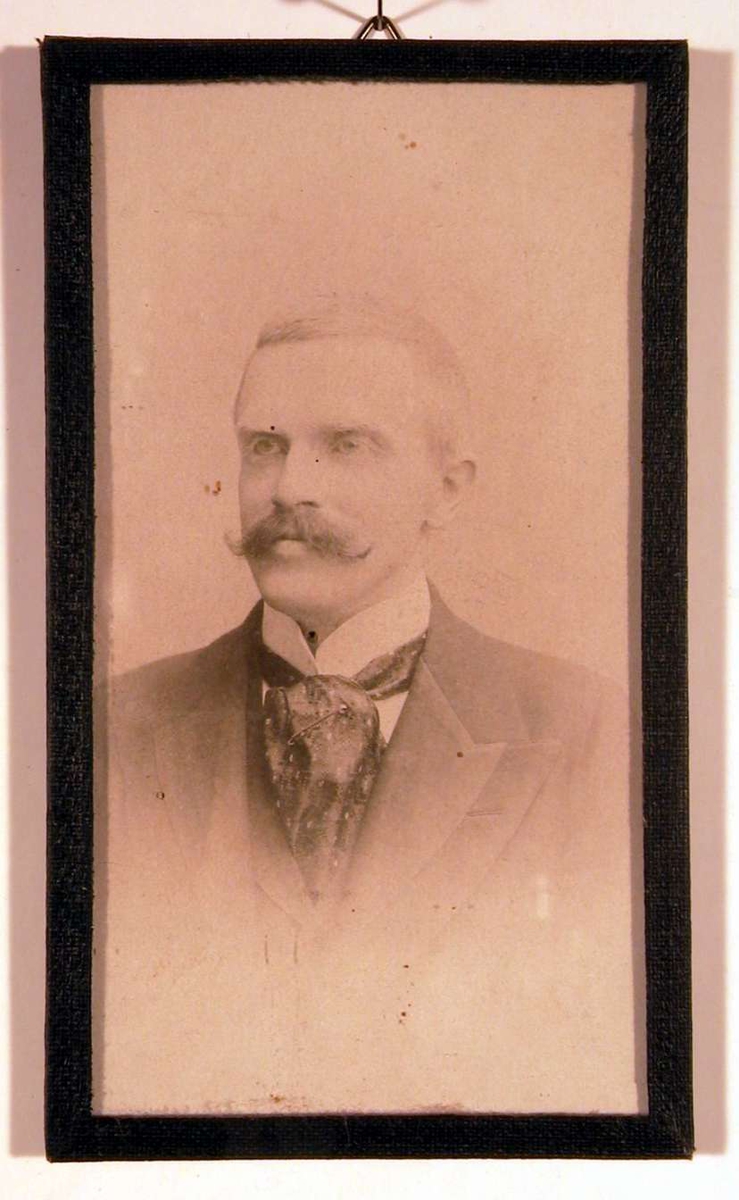 Portrettfotografi av en mann med snurrebart og stort silkesjerf rundt den hvite opprettstående skjortekrave og mørk dress med vest.