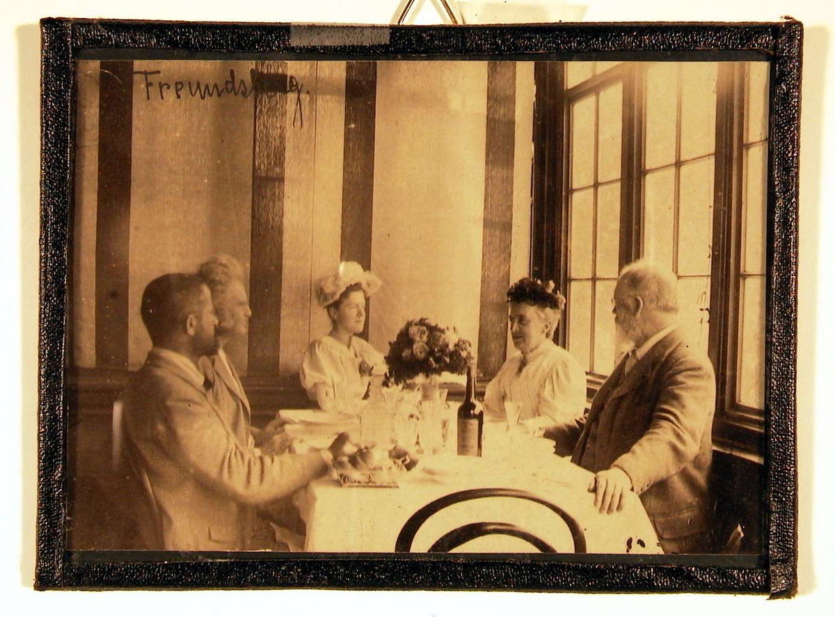 Fotografi fra spisesal; ved bordet sitter to damer og tre herrer; de ser på hverandre.