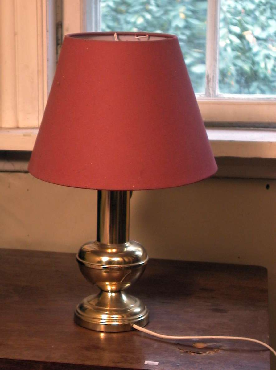 Bordlampe i metall med rød skjerm. Lampen har hvit ledning og støpsel.