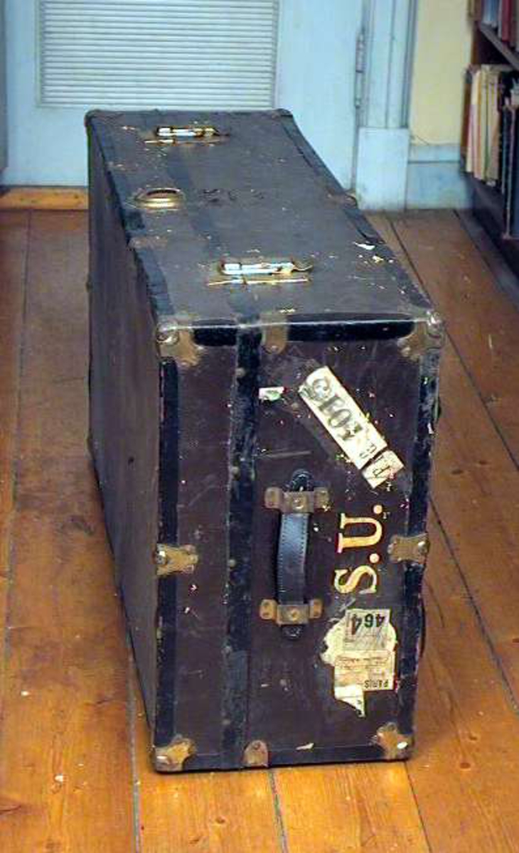 Koffert av finér med sorte metallbeslag (blikk). Kofferten er brunmalt. Den har lærhåndtak på begge sider. Den har lås og beslag av messing. Hovedlåsen er i stykker, men den ligger i kofferten.