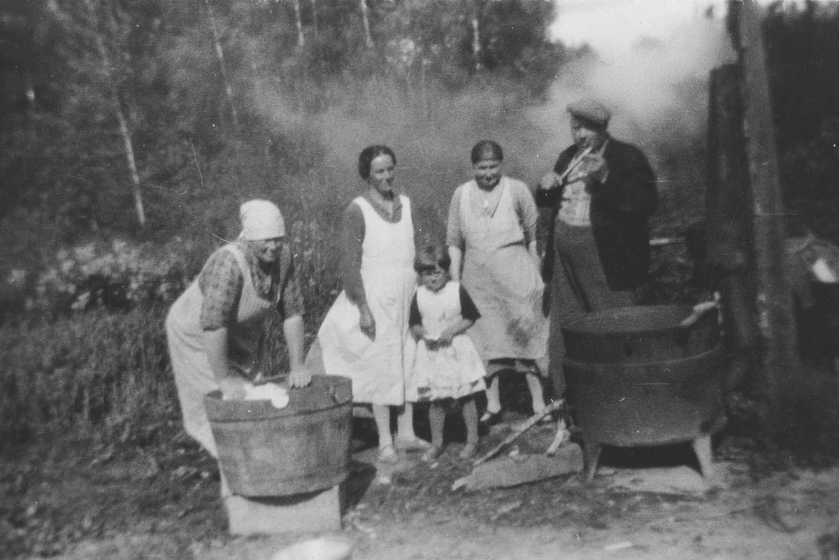 Klesvask utendørs med bryggepanne og stammp. Tre kvinner, en jente og en mann deltar