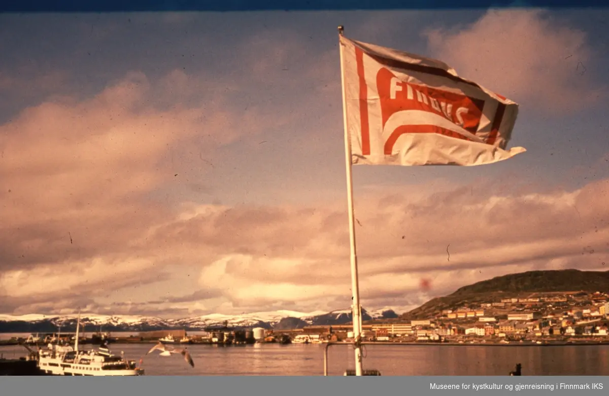Findusflagget vaier over Hammerfest. Fuglenes i bakgrunnen. Ved kaia ligger et hurtigruteskip, sannsynlig "Finnmarken".