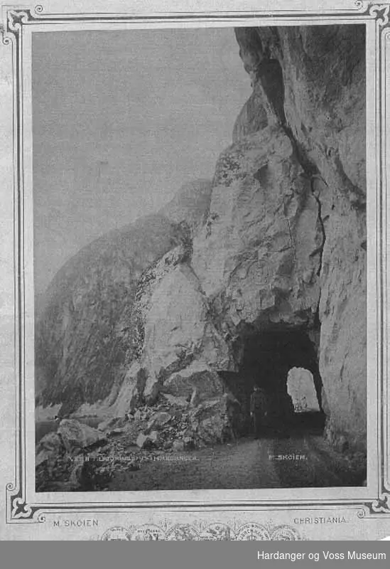 Mann, veg, fjell, vatn. Tunnell langs Eidfjordvatnet med kongevåpenet til Kong Oscar ll