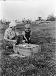 Ukjent kvinne og jente. Sitter ved ei kasse på jordet, leket