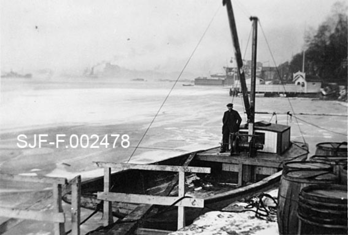 Fraktebåten «Tobias» ved kai på Kongshavn sør for Oslo.  På kaia står en del tretønner på høykant, antakelig tønner som inneholdt tjære- eller bekprodukter som tilhørte firmaet Schwencke & Co, som drev produksjon av slike varer her fram til 1924, da grunnen ble ekspropriert for at jernbanen skulle få plass til dobbeltspor på denne strekningen.  Båten er en liten, bred og flatbotnet farkost med styrhus bakerst og betydelig dekksplass foran.  En mann står ved styrhuset.  Foran ham en slags vinsj, som antakelig ble brukt under lasting og lossing.  I bakgrunnen en vinterkald og delvis islagt Oslofjord.