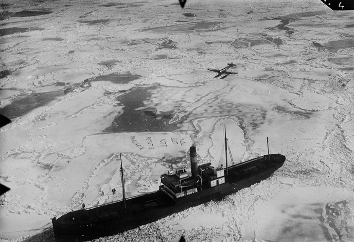 Ett infruset lastfartyg får hjälp av ett överflygande militärt flygplan av typ S 5. På isen har fartygsbesättningen skrivit 'Mjöl, jäst, kött, potatis, margarin'.