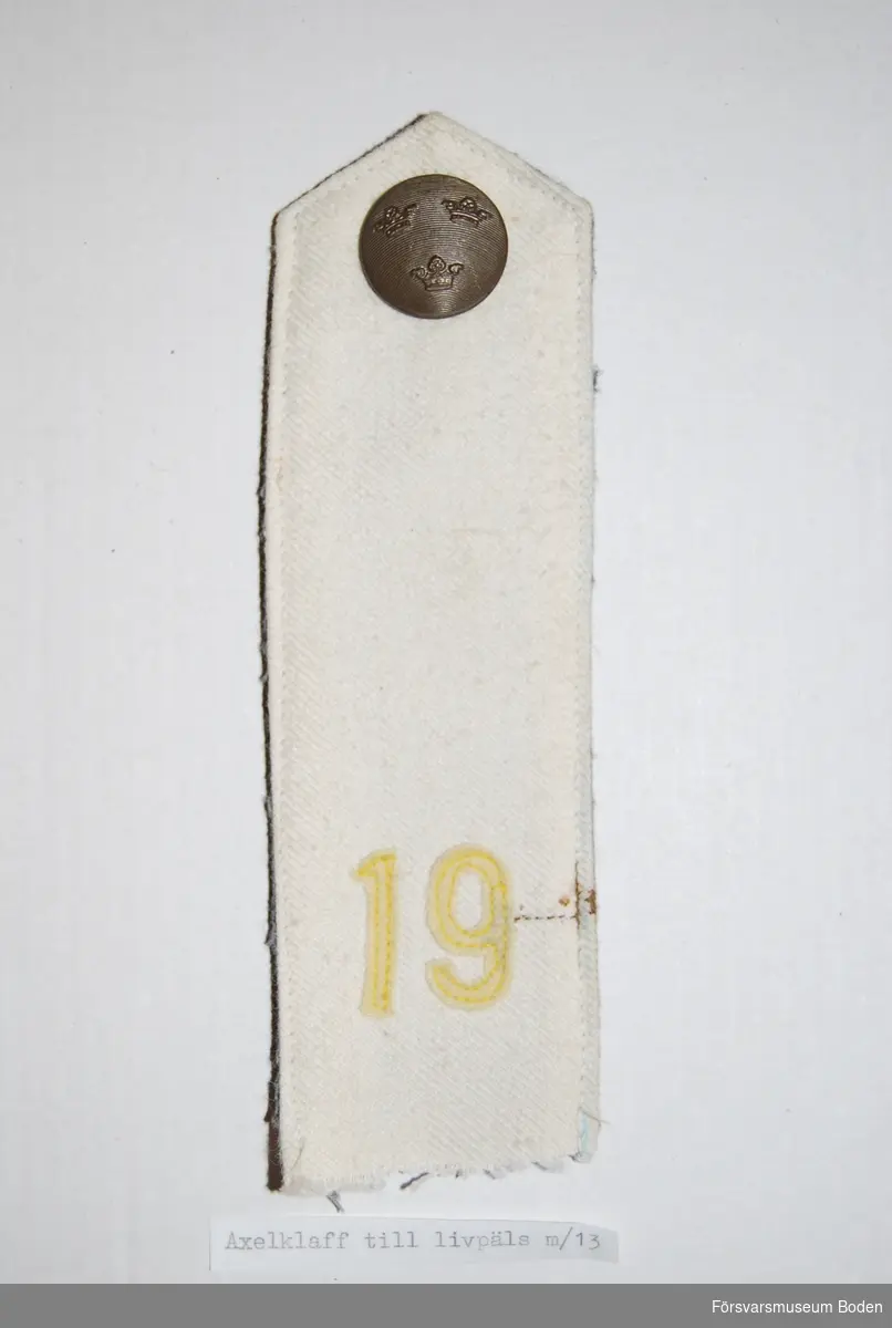 Vitt diagonaltyg med foder av grått kommisskläde. Nertill siffran 19 av gult kläde för Norrbottens regemente (I 19). Högst upp tre kronorknapp i brons. Knapphål på baksidan.
