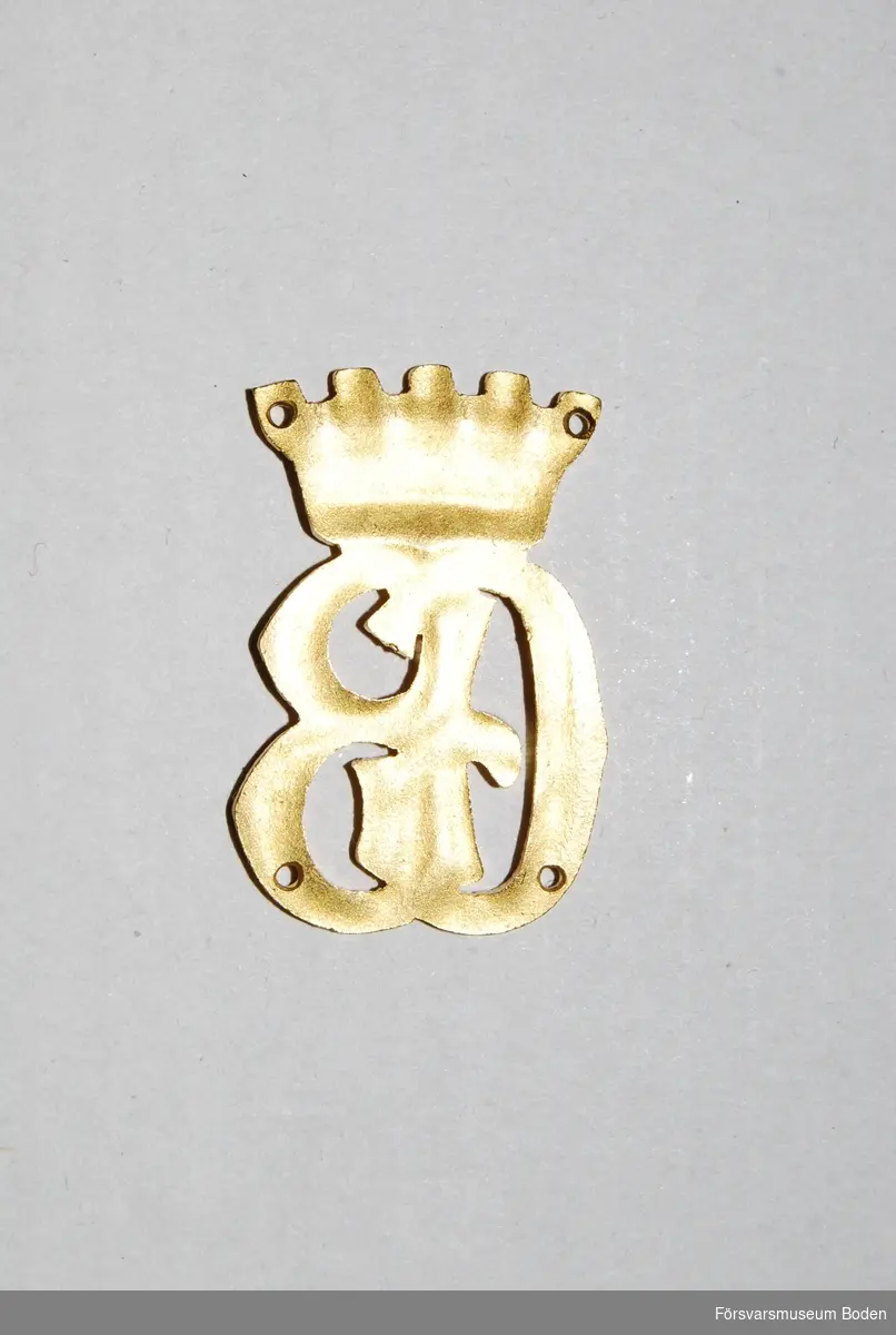 Bokstäverna GB under krona, av guldfärgad metall.