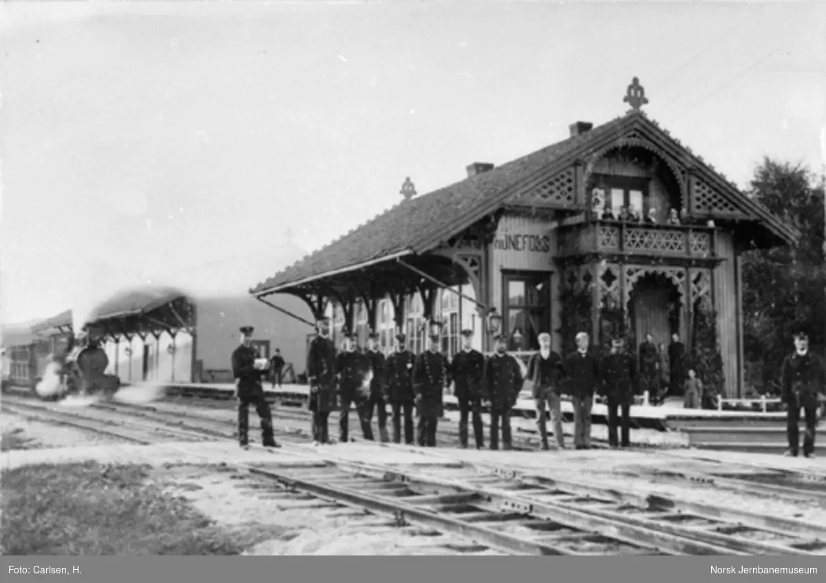 Personalet på Hønefoss stasjon oppstilt for fotografering med stasjonsbygningen og tog 184 "Slaven" i bakgrunnen