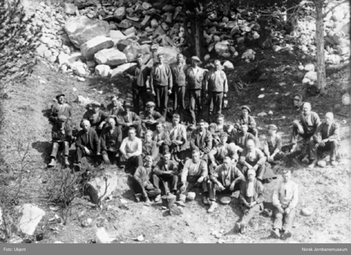 Gruppebilde av 30-40 anleggsarbeidere sittende og stående i en skråning